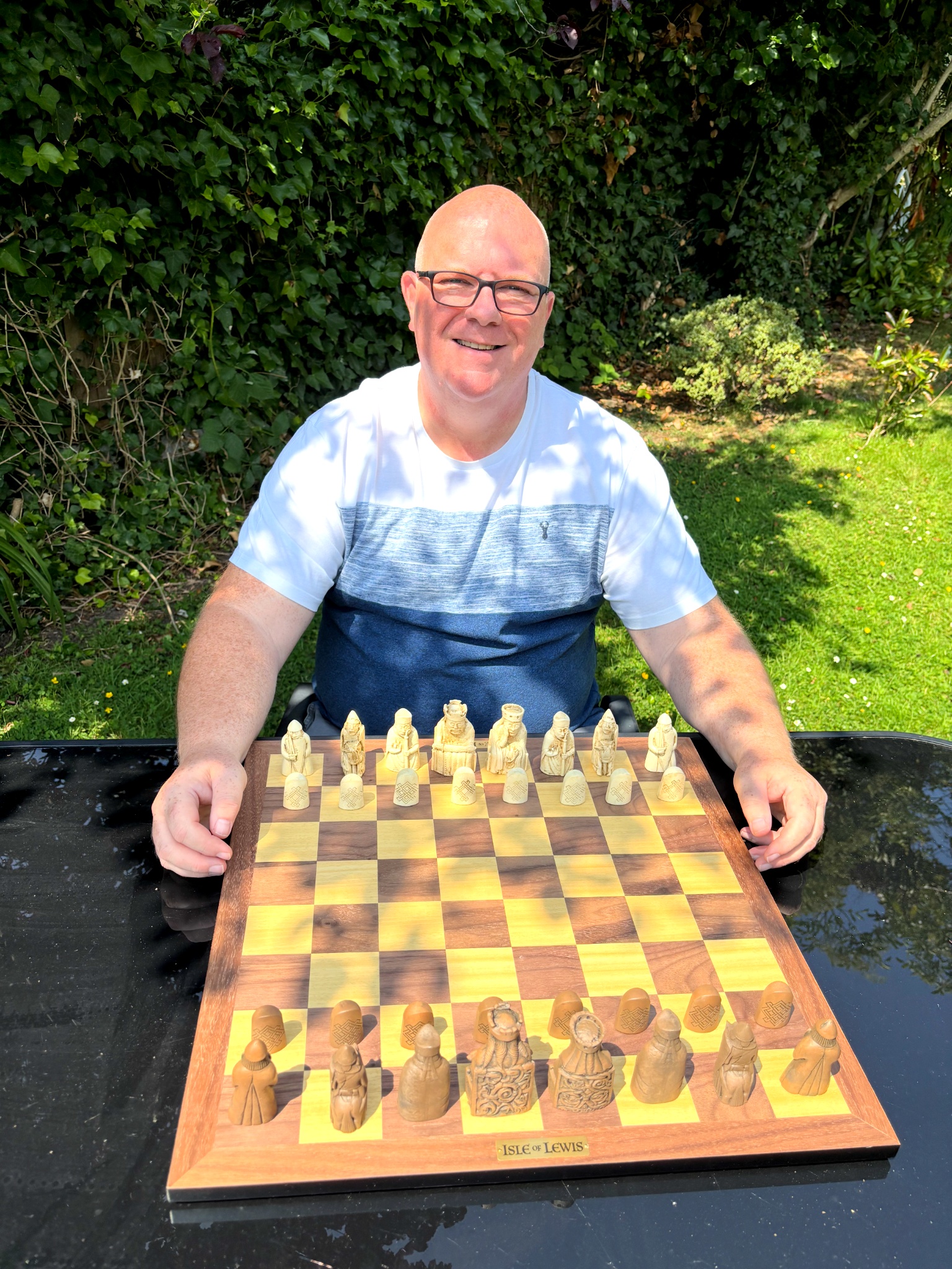 Gregory Hunt seated at an outdoor table with a carved Isle of Lewis chess set, smiling at the camera with the full board set for play in front of him.